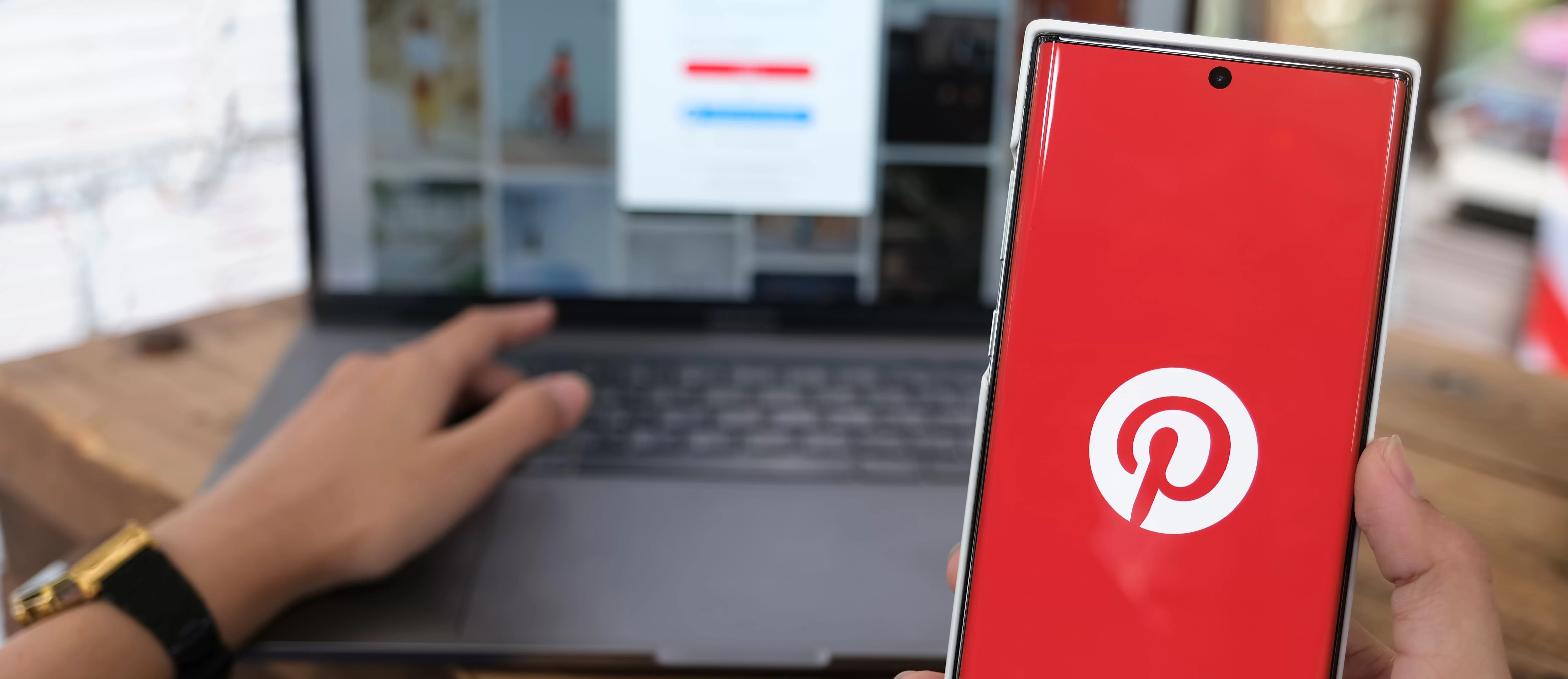 Pessoa segurando um smartphone com a tela exibindo o logotipo do Pinterest em vermelho, enquanto usa um laptop em segundo plano que mostra a interface do Pinterest com imagens e uma tela de login desfocada.