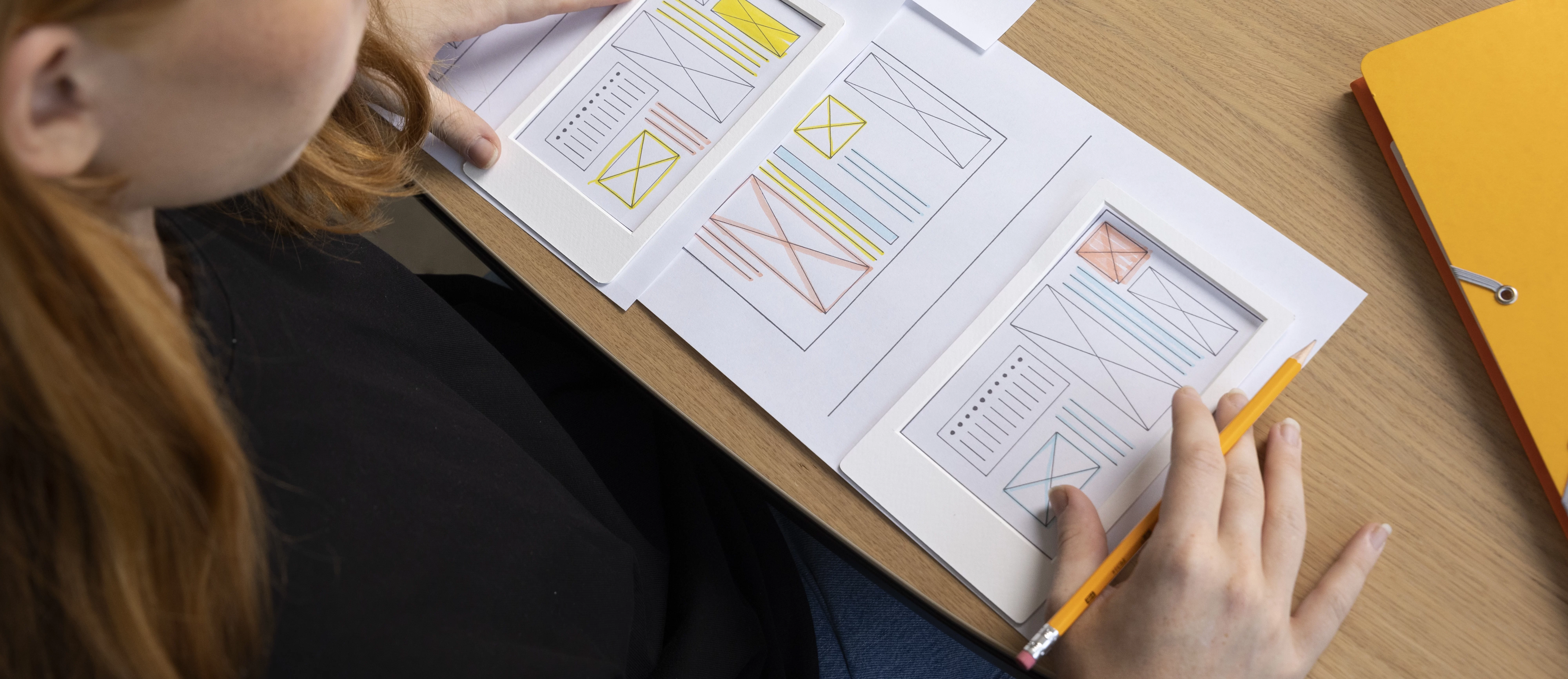 Mulher analisando protótipos de layout de interface de usuário impressos, utilizando moldura para simular tela de dispositivos móveis, durante processo de design e arquitetura da informação em ambiente de trabalho.